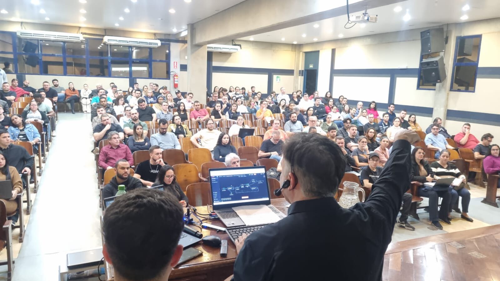 Prof. Dr. Eduardo Mendes gesticulando durante palestra para plateia lotada, com diagrama de automação projetado