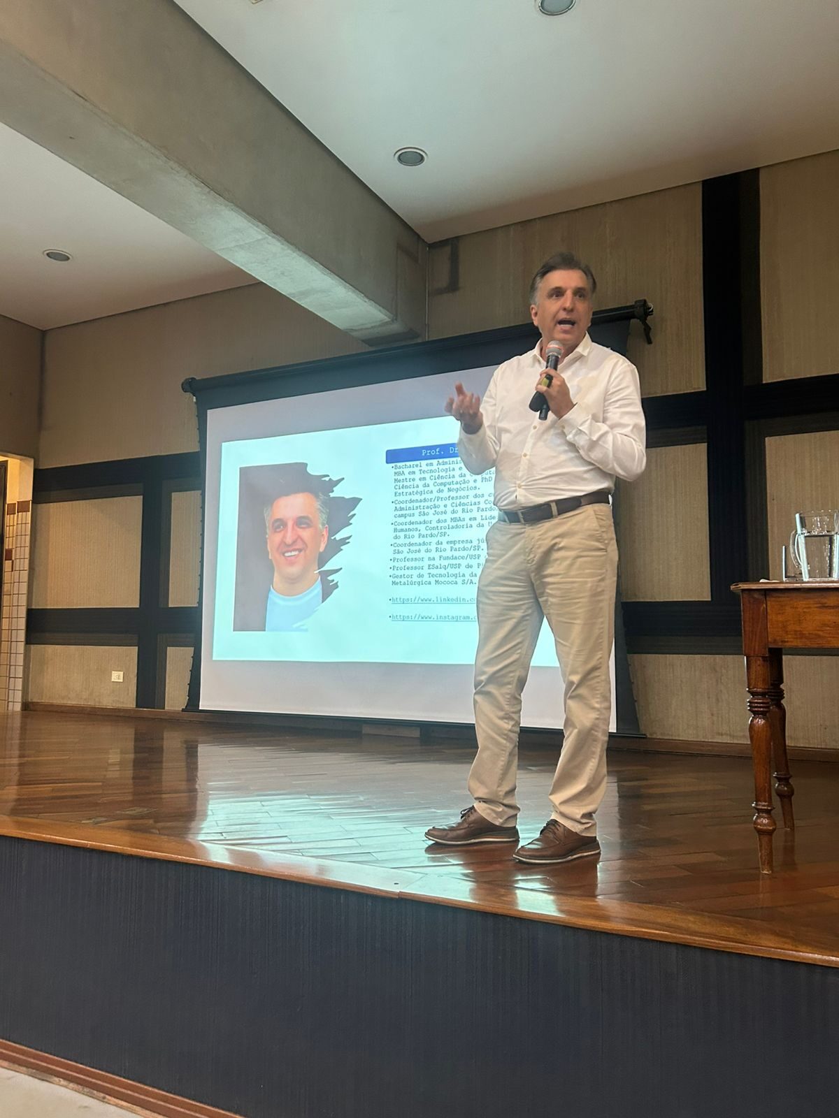 Prof. Dr. Eduardo Mendes apresentando no palco, com microfone, ao lado do slide de sua biografia
